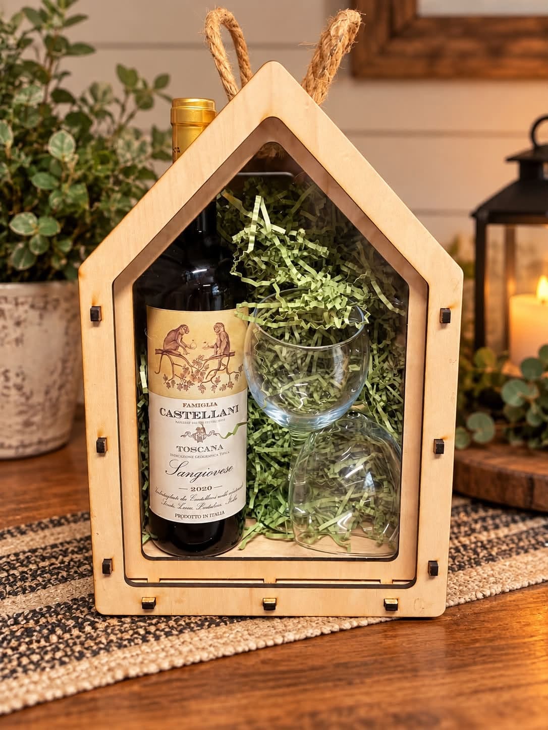 House-shaped maple wine gift box with a clear acrylic sliding lid, shown with a wine bottle, two glasses, and green paper filler.